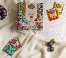 Opakowanie Basilur Vintage Blossoms Assorted ustawione na jasnym materiale w otoczeniu sliwek i pojedynczych torebek herbat o roznych smakach. Kompozycja z kwiatowym motywem na pudełku oraz pastelowymi saszetkami podkresla elegancje i naturalny charakter herbat czarnych i zielonych Basilur.