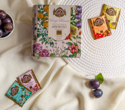 Opakowanie Basilur Vintage Blossoms Assorted ustawione na jasnym materiale w otoczeniu sliwek i pojedynczych torebek herbat o roznych smakach. Kompozycja z kwiatowym motywem na pudełku oraz pastelowymi saszetkami podkresla elegancje i naturalny charakter herbat czarnych i zielonych Basilur.