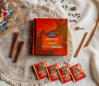 Opakowanie Basilur Breakfast Tea Asia Assorted ustawione na jasnym materialowym tle wsrod ziol i przypraw takich jak cynamon imbir gozdziki oraz galka muszkatolowa. W dolnej czesci widoczne sa czerwone saszetki herbat o roznych wariantach sniadaniowych co podkresla orientalny aromat i bogaty smak czarnej oraz bialej herbaty Basilur.