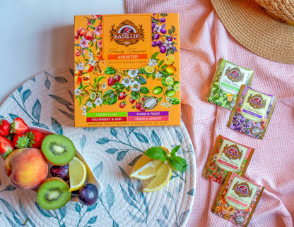 Kolorowe opakowanie Basilur Fruity Fusions Assorted ustawione obok swiezych owocow takich jak truskawki, kiwi, brzoskwinia oraz sliwki. W kadrze widoczne sa takze pojedyncze torebki herbat w roznych smakach ulozone na rozowym materiale co podkresla letni klimat oraz owocowo ziolowy charakter herbat Basilur.