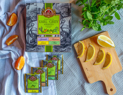 Zestaw herbat Basilur Earl Grey Assorted ustawiony na jasnym materiale z dodatkiem swiezych kawalkow pomaranczy oraz cytryny. W kadrze widac koperteksy roznych smakow oraz galazke miety co podkresla naturalny i aromatyczny charakter zielonej herbaty Earl Grey.