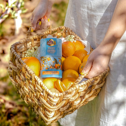 Tuba Basilur Orange and Orange Blossom w wiklinowym koszu pełnym pomarańczy, trzymanym przez osobę w jasnej sukience