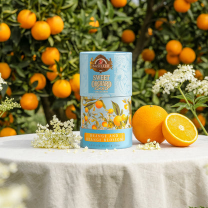 Herbata Basilur Orange and Orange Blossom w tubie, ustawiona przy pomarańczach i białych kwiatach w sadzie