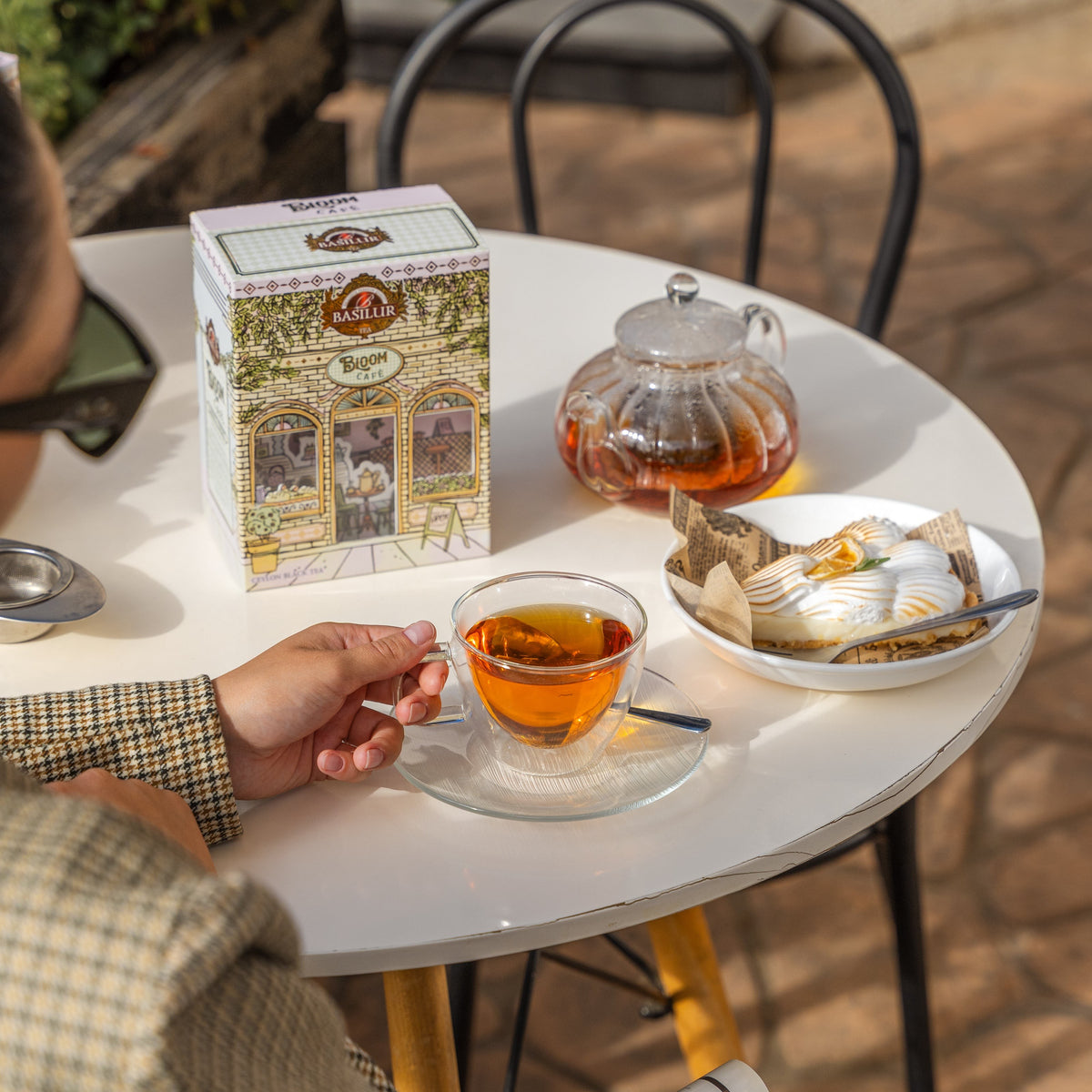 Opakowanie Basilur Bloom Cafe na stoliku obok filiżanki z herbatą, szklanego dzbanka i deseru