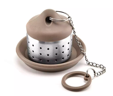 Brązowy zaparzacz do herbaty na podstawce z łańcuszkiem i uchwytem.