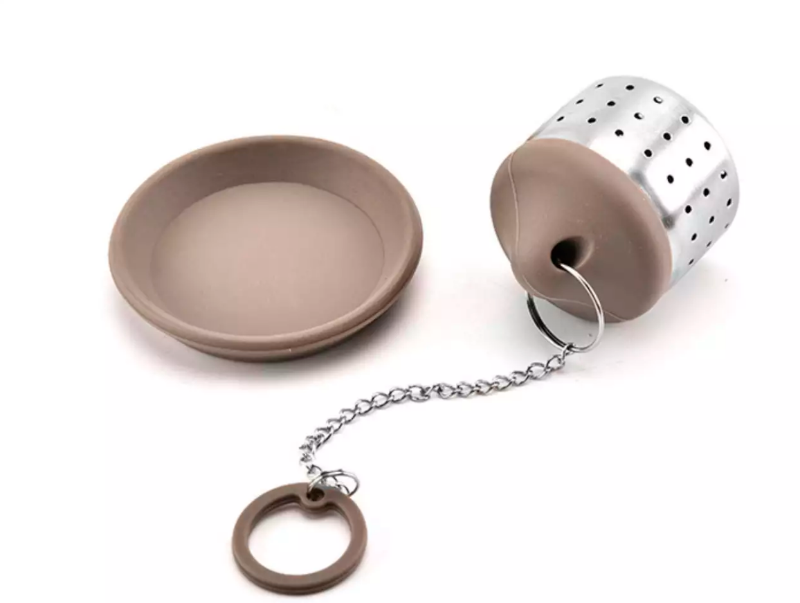 Zaparzacz do herbaty w kolorze brązowym z łańcuszkiem i podstawką obok.