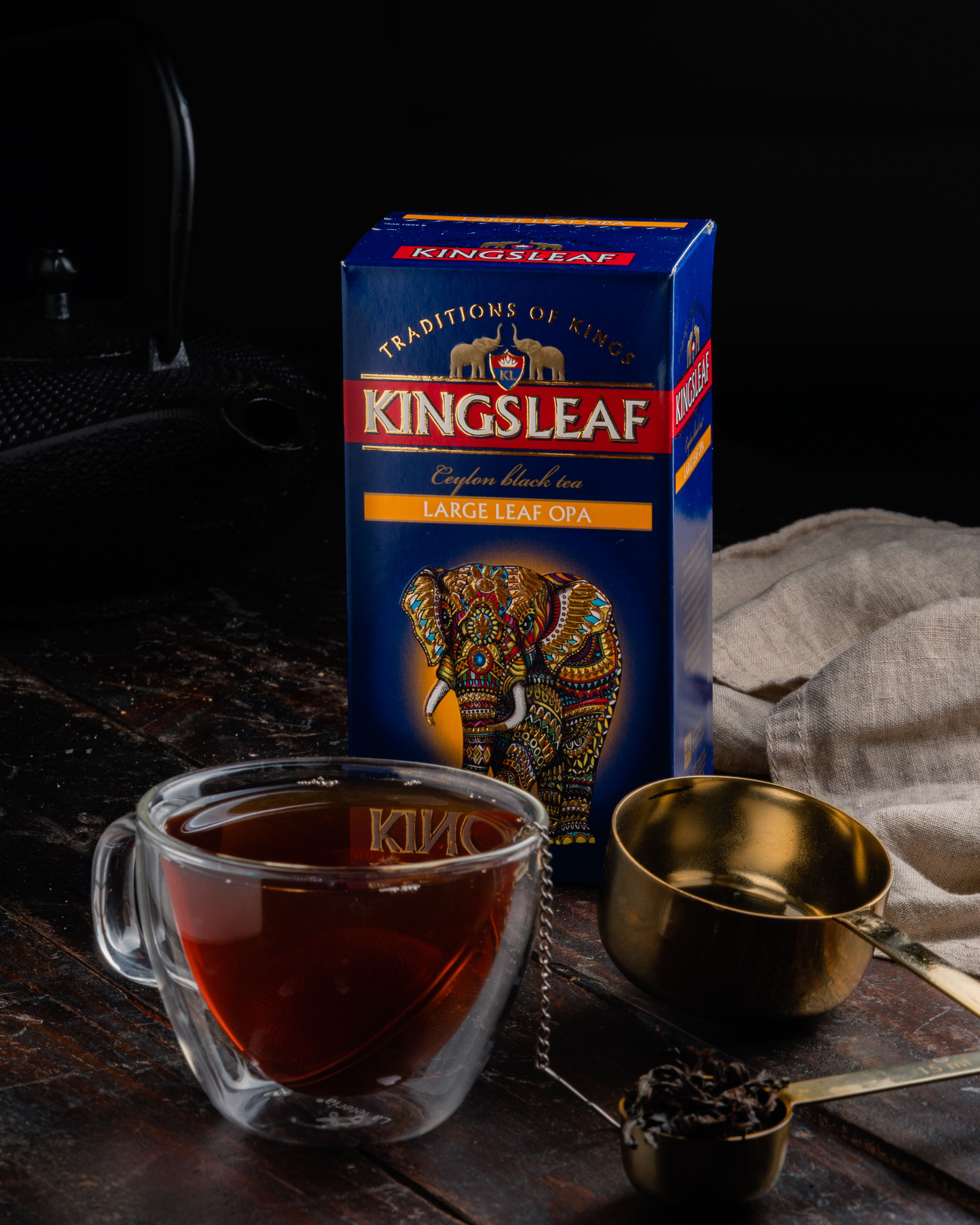 Pudełko herbaty Kingsleaf Large Leaf OPA obok filiżanki z ciemnym naparem i złotej łyżki.