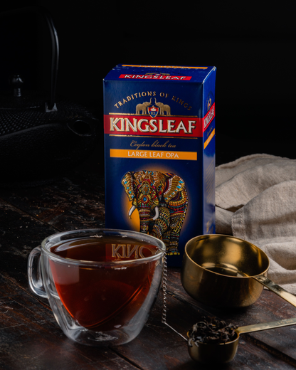 Pudełko herbaty Kingsleaf Large Leaf OPA obok filiżanki z ciemnym naparem i złotej łyżki.