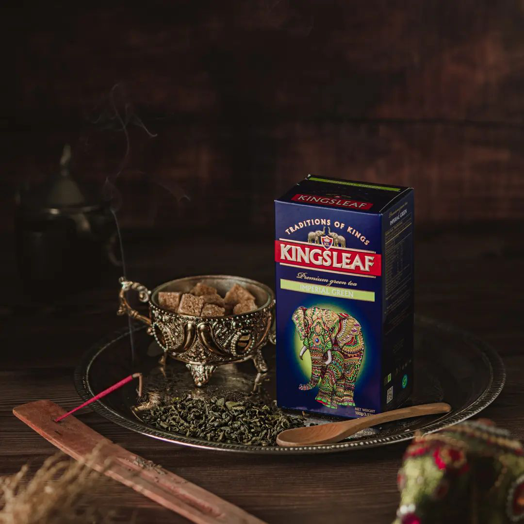 Opakowanie herbaty Kingsleaf Imperial Green na ciemnym tle z liśćmi zielonej herbaty – elegancka kompozycja produktu.