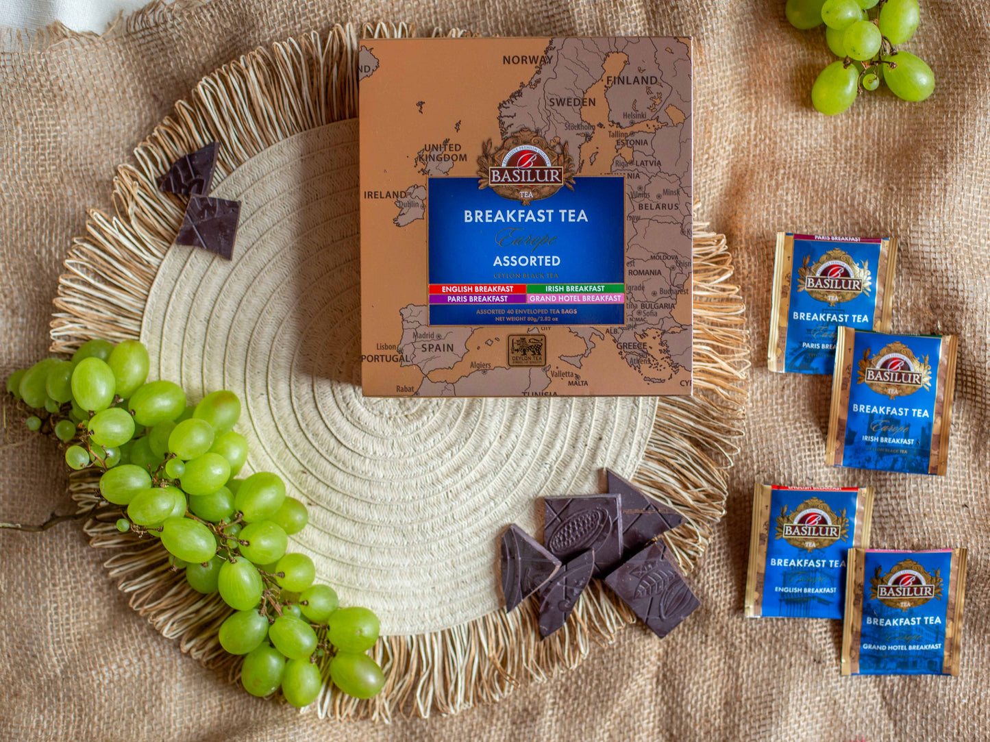 Opakowanie Basilur Breakfast Tea Europe Assorted ustawione na naturalnym jutowym tle w otoczeniu zielonych winogron oraz kostek gorzkiej czekolady. Obok znajduja sie saszetki herbat w wariantach europejskich takich jak English Breakfast Irish Breakfast Paris Breakfast oraz Grand Hotel Breakfast co podkresla klasyczny sniadaniowy charakter czarnej herbaty Ceylon.