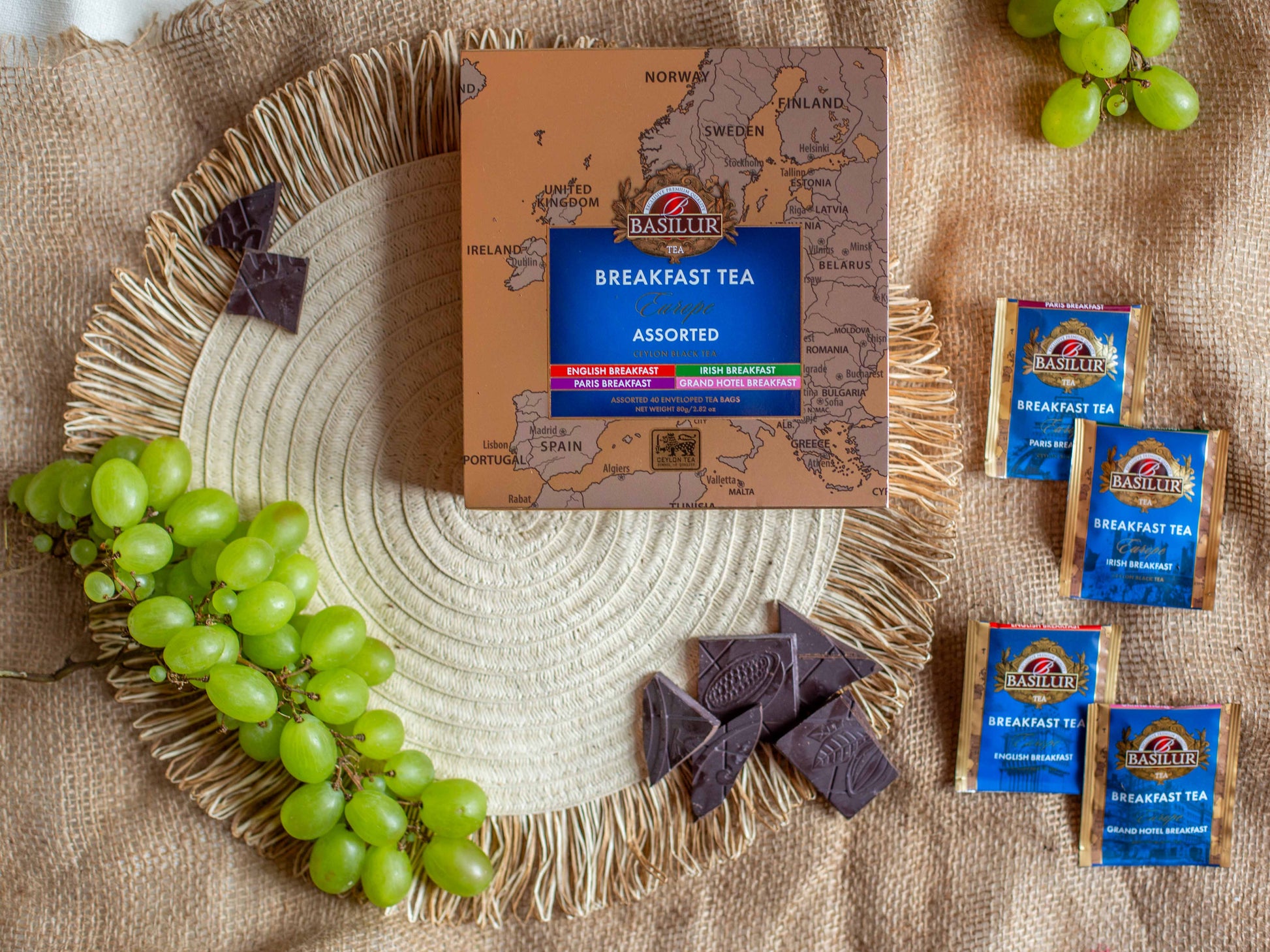 Opakowanie Basilur Breakfast Tea Europe Assorted ustawione na naturalnym jutowym tle w otoczeniu zielonych winogron oraz kostek gorzkiej czekolady. Obok znajduja sie saszetki herbat w wariantach europejskich takich jak English Breakfast Irish Breakfast Paris Breakfast oraz Grand Hotel Breakfast co podkresla klasyczny sniadaniowy charakter czarnej herbaty Ceylon.