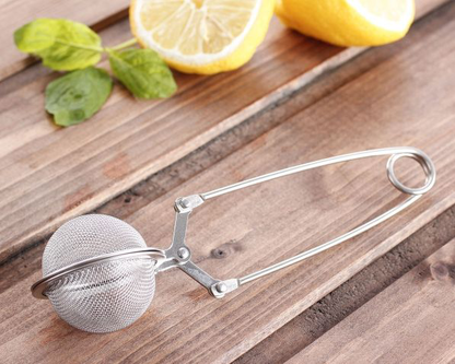 Zaparzacz do herbaty ze stali nierdzewnej w formie szczypiec leżący na drewnianym stole obok cytryn i mięty