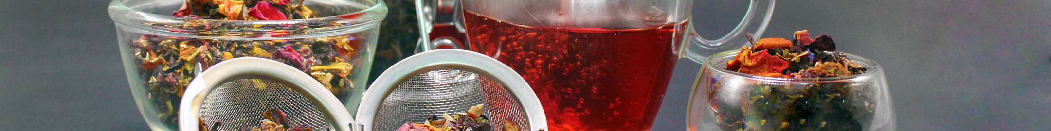 Szklane filiżanki i dzbanek z zaparzoną herbatą ziołową oraz zaparzacze z suszem herbacianym w tle.