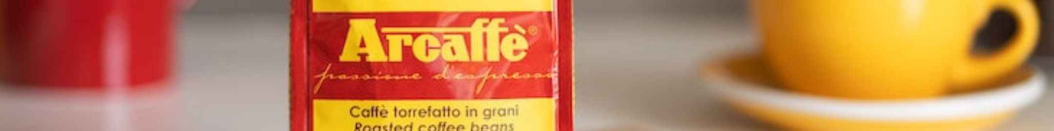 Opakowanie kawy Arcaffè w czerwono-żółtej szacie graficznej z napisem passione d’espresso, ustawione obok filiżanki espresso na tle kawowej scenerii.