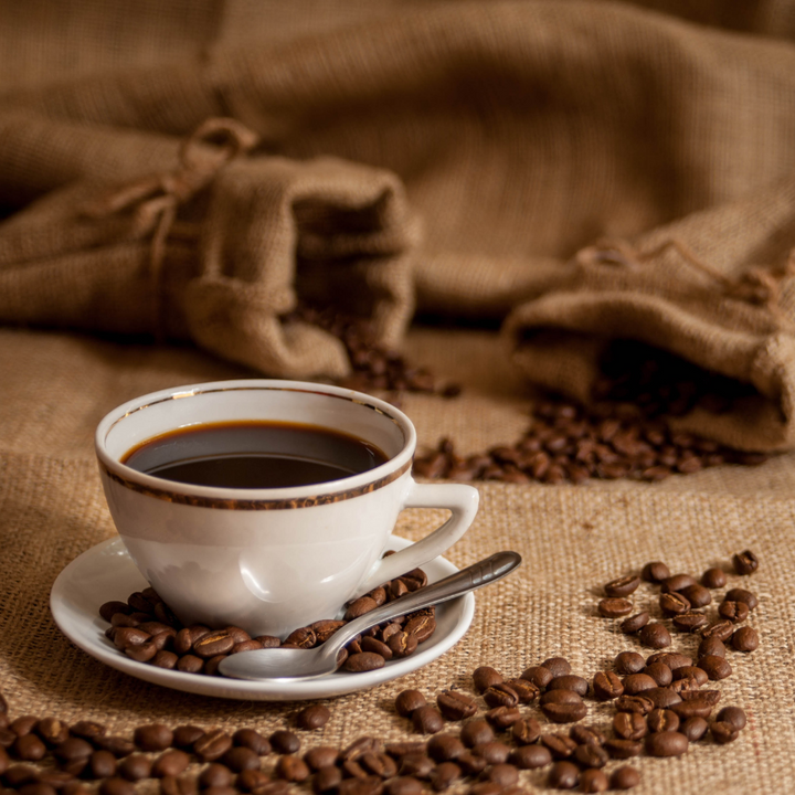 White cup of coffee with a spoon on a saucer surrounded by scattered coffee beans on a rustic fabric background.