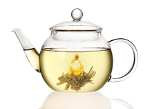 Szklany dzbanek z jasnym naparem białej herbaty oraz rozwiniętym kwiatem herbaty rozkwitającej Autumn Beauty.