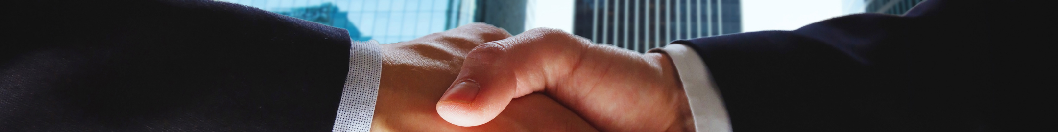 Uścisk dłoni dwóch osób w garniturach na tle nowoczesnych biurowców – symbol partnerstwa i współpracy biznesowej.