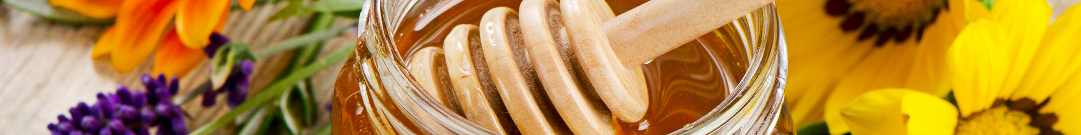 Słoik miodu z drewnianym nabierakiem, otoczony świeżymi kwiatami.