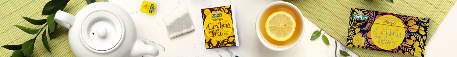 Herbata Stassen Mango Ceylon Tea w torebkach – opakowanie, filiżanka herbaty z plasterkiem cytryny i biały czajniczek na tle bambusowej maty.
