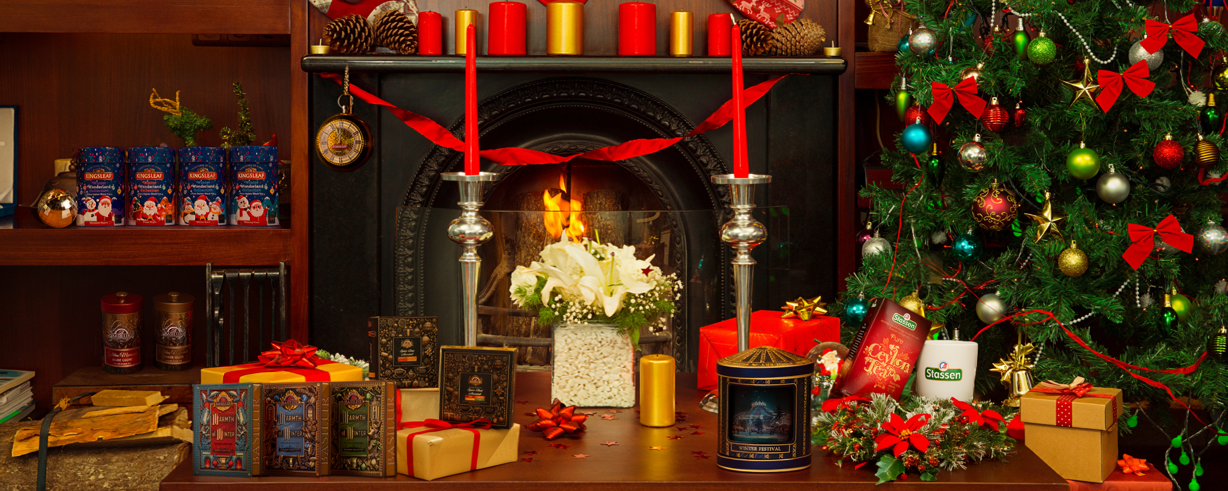 Świąteczna aranżacja z kominkiem, ozdobami, choinką i zestawami herbat ustawionymi wśród prezentów i dekoracji.